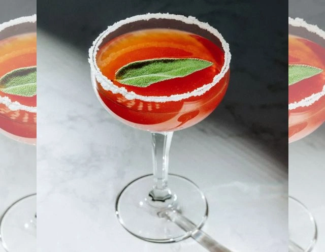 Negroni in mid-century modern glass with beveled edge square and rectangle design - red/orange liquid getting darker toward bottom of glss, with ice and lmon garnish - on round table cover made of sparkly silver beeds and round copper, grey, taupe, and silver small circle coins around edges.