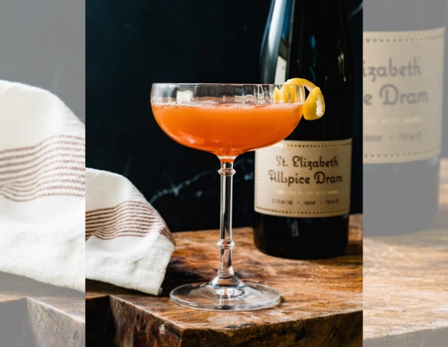 Negroni in mid-century modern glass with beveled edge square and rectangle design - red/orange liquid getting darker toward bottom of glss, with ice and lmon garnish - on round table cover made of sparkly silver beeds and round copper, grey, taupe, and silver small circle coins around edges.